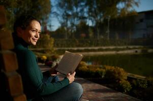 portret van een tevreden mooi vrouw zittend Aan houten bank in park, lezing boek, genieten van warm zonnig herfst dag , weg van de drukte en drukte van stad, resting van digitaal gadgets en werk foto