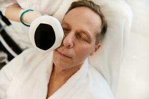 detailopname van een knap Europese Mens ontspannende in welzijn spa gedurende mesotherapie Cursus met modern medisch uitrusting voor gezicht aanscherping Bij schoonheidsspecialiste kliniek foto