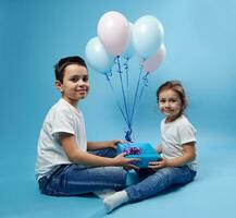 jongen en meisje gekleed gelijk zitten Aan blauw achtergrond en houden een blauw geschenk met roze boog en kijken Bij camera foto