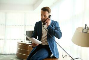 attent verbaasd jong bedrijf investeerder, zakenman, financiën regisseur zittend Aan hotel tafel hoek met documenten en contract ik handen en pratend ernstig Aan vaste telefoon telefoon gedurende bedrijf reis foto