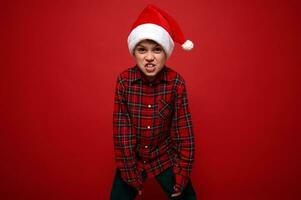 grappig preadolescent jongen in plaid overhemd en de kerstman claus hoed grimassen op zoek Bij camera poseren tegen rood gekleurde achtergrond met kopiëren ruimte voor Kerstmis en nieuw jaar advertentie foto