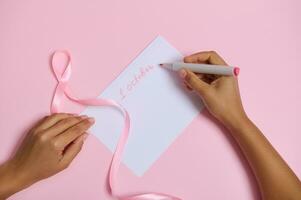 vlak leggen van vrouw hand- Holding voelde pen, schrijft 1 oktober Aan wit blanco vel van papier, roze lint symbool van oktober borst kanker bewustzijn maand, aan het liegen Aan roze achtergrond met ruimte voor tekst foto