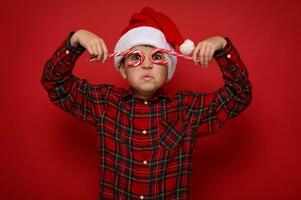 aanbiddelijk preteen jongen in de kerstman claus hoed en plaid overhemd looks Bij camera door Kerstmis zoet snoep wandelstokken, Holding hen imiteren bril, poseren Aan rood achtergrond met kopiëren ruimte voor advertentie foto