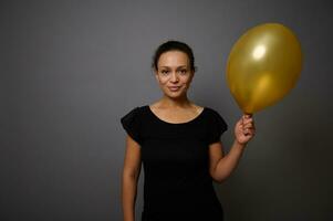 vrolijk Afrikaanse Amerikaans vrouw gekleed in zwart glimlacht op zoek Bij camera en poses tegen grijs achtergrond met een goud gekleurde lucht ballon in hand. zwart vrijdag concept met kopiëren ruimte voor advertentie foto