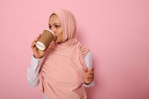mooi Arabisch moslim vrouw met gedekt hoofd drinken heet drankje, thee of koffie van beschikbaar karton meenemen beker, staand zijwaarts, drie kwartalen, tegen roze achtergrond met kopiëren ruimte foto