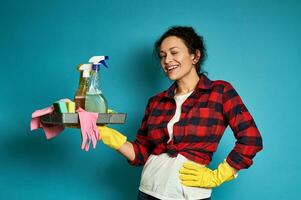 glimlachen vrouw poseren Aan blauw achtergrond met dienblad met schoonmaak producten in handen en een hand- Aan taille foto