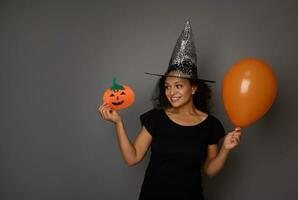 jong mooi vrouw vervelend een tovenaar hoed, gekleed in zwart kleding, Holding een kleurrijk oranje lucht ballonnen en shows een vilt gesneden handgemaakt pompoen, glimlacht met mooi toothy glimlach. halloween concept foto