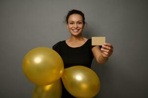 mooi glimlachen gelukkig vrouw gekleed in zwart kleding shows opgeblazen lucht ballonnen van goud kleur en goud leeg blanco credit kaart. zwart vrijdag concept met kopiëren advertentie ruimte Aan grijs muur achtergrond foto