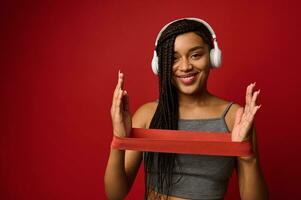 mooi vrouw van Afrikaanse etniciteit met elegant dreadlocks gebruik makend van rood elastisch geschiktheid band terwijl aan het doen uitrekken opdrachten Aan armen, glimlacht toothy glimlach, geïsoleerd Aan gekleurde achtergrond met kopiëren ruimte foto