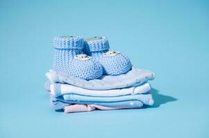 schattig blauw gebreid baby laarzen Aan een stapel van schoon witgewassen gestreken pasgeboren bodysuits, geïsoleerd over- blauw pastel achtergrond foto