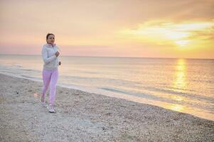 vol lengte aantrekkelijk atletisch vrouw aan het doen ochtend- joggen langs de kustlijn van de zee tegen de backdrop van een mooi zonsopkomst foto