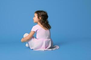 visie van de terug van een meisje ballerina gekleed in roze uniform op zoek weg zittend Aan blauw achtergrond met kopiëren ruimte foto