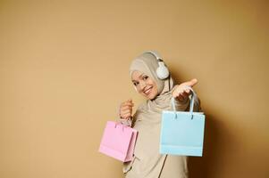 aanbiddelijk moslim vrouw in hijab met een mooi toothy glimlach staat zijwaarts naar de camera en poseren Holding uit een geschenk zak Aan een beige achtergrond met kopiëren ruimte foto