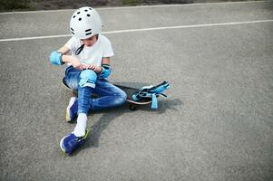 voorkant visie van een sportief jongen in veiligheid helm zittend Aan skateboard en zetten Aan beschermend knie stootkussens foto