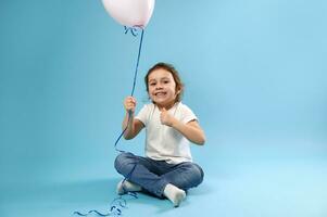 charmant weinig meisje met een roze ballon in haar hand- tonen een duim omhoog Aan een blauw achtergrond met kopiëren ruimte voor Promotie en reclame. foto