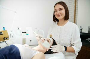 jong schoonheidsspecialist dokter toepassen schoonheid hydraterend masker naar gezicht van blond vrouw aan het liegen Aan massage tafel in schoonheid spa kliniek. professioneel huid zorg concept foto