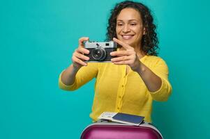 aantrekkelijk midden volwassen spaans vrouw in helder geel trui fotograferen met een wijnoogst camera, glimlachen toothy glimlach poseren met koffer, instappen voorbij gaan aan en ticket Aan blauw achtergrond. kopiëren ruimte foto