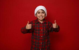 knap preadolescent jongen in rood geruit overhemd vervelend een de kerstman hoed glimlacht toothy glimlach en shows duimen omhoog op zoek Bij camera poseren tegen gekleurde achtergrond met kopiëren ruimte voor Kerstmis advertentie foto