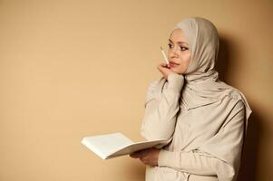 aantrekkelijk jong vrouw in een hijab zijwaarts naar de camera met een Open dagboek in een hand- en een potlood in de andere in de buurt haar kin en looks peinzend naar de kant foto