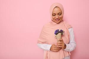 geïsoleerd zelfverzekerd portret Aan gekleurde achtergrond met kopiëren ruimte van een aantrekkelijk moslim Arabisch vrouw in roze hijaab, op zoek Bij een schattig boeket van wilde bloemen in Purper tinten, verpakt in ambacht papier foto