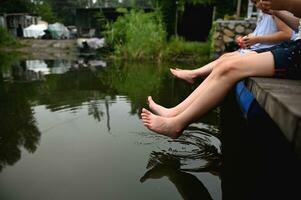 detailopname van een jongen en een meisje, broer en zus zijn zittend Aan de pier Aan een warm zomer dag, bungelend hun voeten in de water en spatten in de voorgrond foto