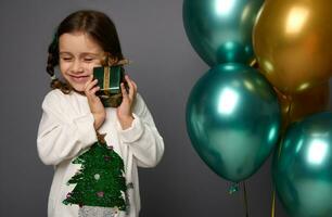 geïsoleerd portret Aan grijs achtergrond van aanbiddelijk Kaukasisch weinig meisje met Gesloten ogen voorzichtig knuffelen haar Kerstmis geschenk, staand in de buurt mooi glimmend lucht ballen van groen metalen en gouden kleuren foto