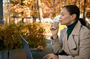 mooi vrouw genieten van ver weg werk, chillen in de houten cafetaria in de eik bosje en drinken heerlijk heet koffie terwijl werken Aan laptop weg van de drukte en drukte van de stad foto