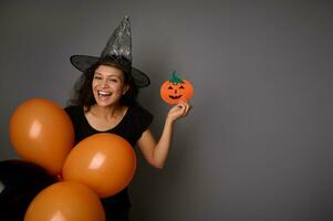 mooi vrouw in tovenaar hoed, gekleed in zwart, houdt oranje ballonnen en handgemaakt vilt gesneden zwart pompoen, glimlacht op zoek Bij camera, poses over- grijs achtergrond, kopiëren ruimte . halloween partij concept foto