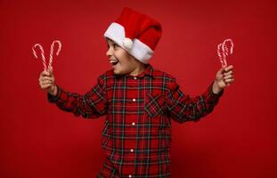 aanbiddelijk kind jongen in rood plaid overhemd en de kerstman claus hoed op zoek Bij Kerstmis lolly, gestreept suikerachtig snoep wandelstokken in zijn handen, geïsoleerd Aan rood gekleurde achtergrond met kopiëren ruimte voor advertentie foto