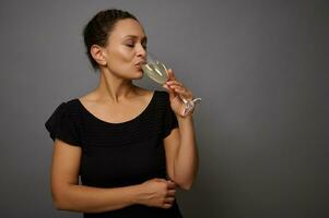 vrolijk vrouw in zwart kleding drankjes sprankelend wijn van een glas, staand geïsoleerd over- grijs muur achtergrond met kopiëren advertentie ruimte foto