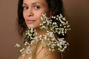 mooi vrouw met wit gypsophila takje staand drie kwart tegen bruin achtergrond op zoek vol vertrouwen Bij camera. lichaam zorg, positiviteit, vrouwelijkheid, vrouwen dag . detailopname portret. foto