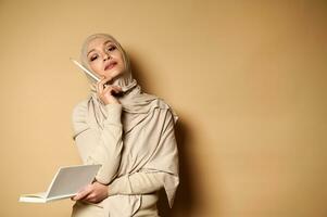 aantrekkelijk Arabisch moslim vrouw in hijab houdt een dagboek en looks omhoog bedachtzaam, richten met een wit potlood Aan een beige achtergrond met kopiëren ruimte. foto