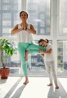 jong vrouw staand in boom houding terwijl beoefenen yoga, haar dochter resting haar hoofd Aan haar knie en glimlachen schattig Bij camera foto