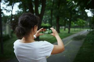 achterzijde visie van een jong vrouw Holding mobiel telefoon in leven visie mode camera, nemen foto van de park steeg