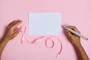 vlak leggen van vrouw hand- Holding voelde pen klaar naar schrijven Aan wit blanco vel van papier, roze lint symbool van oktober borst kanker bewustzijn maand, aan het liegen Aan roze achtergrond met ruimte voor tekst foto