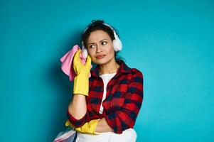 aantrekkelijk vrouw, huisvrouw, staand tegen blauw achtergrond met schoonmaak gereedschap en benodigdheden, op zoek aandachtig, met een nadenkend kijken foto