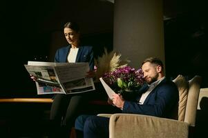 twee knap mensen, midden- oud Kaukasisch Mens en mooi elegant vrouw in bedrijf pak, collega's, bedrijf partners lezing een krant- in de hotel lobby foto