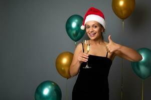 aanbiddelijk vrouw in een de kerstman hoed shows een fluit met Champagne en duim omhoog, glimlacht op zoek Bij camera tegen Kerstmis decor met luxe glimmend lucht ballonnen Aan grijs muur achtergrond met kopiëren advertentie ruimte foto