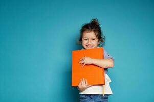 aanbiddelijk weinig meisje voorzichtig knuffels een oranje boek en schattig glimlacht met toothy glimlach op zoek Bij camera terwijl staand Aan blauw achtergrond met kopiëren ruimte foto