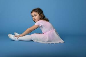 kant portret van een weinig ballerina in roze jurk het uitvoeren van uitrekken poten, zittend over- blauw achtergrond foto