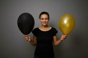 vrolijk Afrikaanse Amerikaans vrouw gekleed in zwart glimlacht op zoek Bij camera en poses tegen grijs achtergrond met een goud en zwart lucht ballonnen in hand. zwart vrijdag concept met kopiëren advertentie ruimte foto