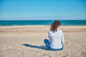 achterzijde visie van een jong vrouw in wit overhemd en blauw jeans zittend Aan de zand in de strand Aan een mooi zonnig dag foto