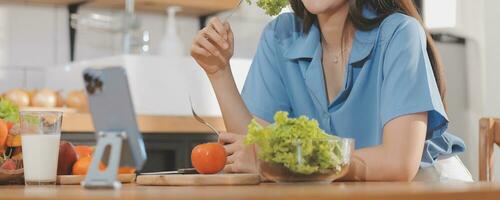 een jong vrouw met een mooi gezicht in een blauw overhemd met lang haar- aan het eten fruit zittend binnen de keuken Bij huis met een laptop en notitieboekje voor ontspanning, concept vakantie. foto