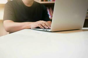 beeld van een jong Mens werken Aan zijn laptop in bibliotheek, achterzijde visie van bedrijf Mens handen bezig gebruik makend van laptop Bij kantoor bureau, jong mannetje leerling typen Aan computer zittend Bij houten tafel foto