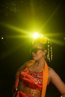 een Sundanees danser poses in voorkant van een schijnend licht terwijl vervelend een oranje kostuum met zonnebril en bloemen Aan haar hoofd foto