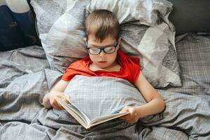 jongen met bril leugens Aan een bed onder een deken en leest een boek, top visie. een kind van primair school- leeftijd is lezing een boek in zijn bed. foto