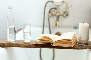 een boek, een fles van water, een glas en een kaars Aan een houten dienblad bovenstaand de badkamer, tegen de achtergrond van een antiek kraan foto