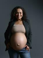 mooi Afrikaanse Amerikaans vrouw, verwachtend moeder glimlachen Bij camera, poseren kaal zwanger buik over- grijs achtergrond foto