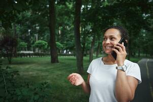 charmant multi-etnisch jong vrouw pratend Aan mobiel telefoon terwijl wandelen de steeg van een stad park Aan een zomer dag foto
