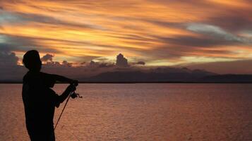 silhouet van een visser met een visvangst hengel in de meer Bij zonsondergang foto